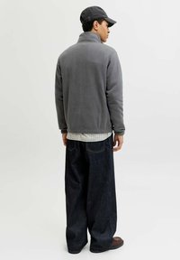 Veste en polaire grise avec col montant, portée sur une chemise à carreaux. Pantalons en denim foncé amples et chaussures marron, avec une casquette grise portée à l'envers.