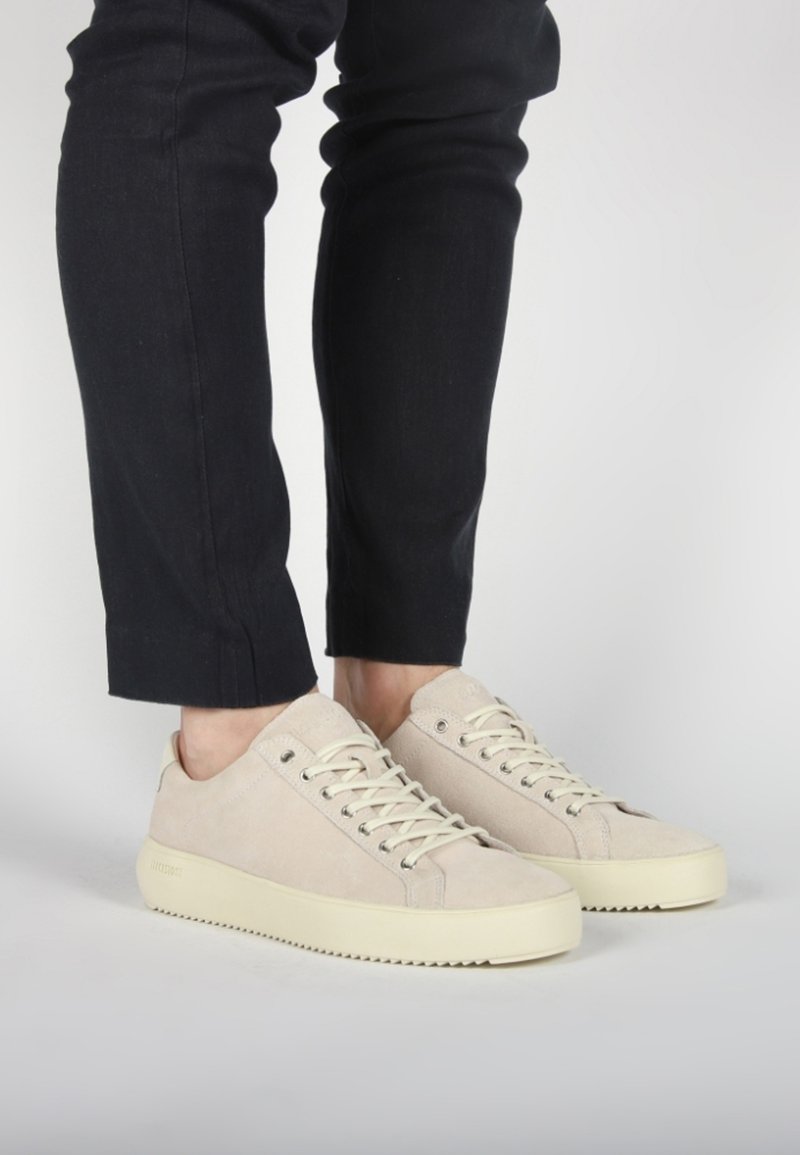 Beige suede sneakers with a textured surface, flat laces, and a thick cream rubber sole, paired with dark slim-fit pants.