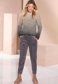 Pullover in einem Farbverlauf von hell- zu dunkelgrau, aus weichem Material. Graue Hose mit seitlichen Reißverschlüssen und hellbraunen Stiefeletten. Minimalistischer Hintergrund.