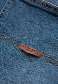 Tissu en denim bleu moyen avec une texture visible. Une étiquette en cuir marron porte l'inscription « ombre dept. depuis 1986 » cousue sur une couture.