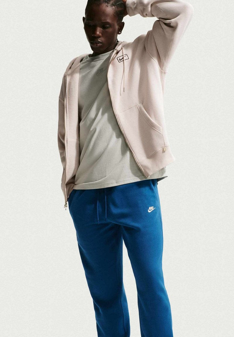 Ljusrosa hoodie med dragkedja över en grå tröja, kombinerad med blåa mjukisbyxor med en vit Nike-logotyp. Slät tyg, avslappnad passform.