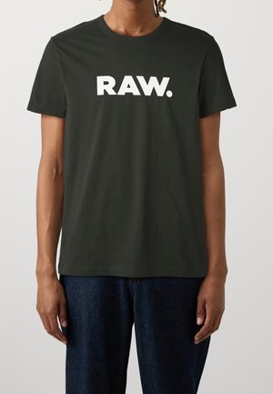 Person iført en sort t-shirt med korte ærmer med "RAW." trykt med store hvide bogstaver foran, kombineret med mørkeblå jeans.