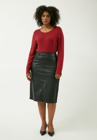 Roter gestrickter Pullover mit Rautenmuster, kombiniert mit einem schwarzen Leder-Midirock mit einem Gürtel. Das Outfit wird durch schwarze High Heels ergänzt.