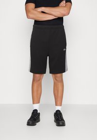 Shorts de sport noirs avec panneaux latéraux gris, taille élastique et logo. Portés avec des baskets noires et des chaussettes blanches.