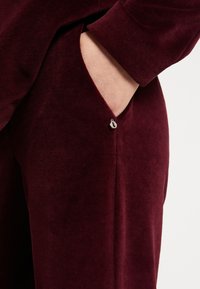 Bordo tkanina s glatkom teksturom, koja ima bočni džep ruban kontrastnim šavom i malim srebrnim dugmetom kao detaljem.