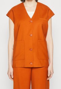 Oranžová bezrukávová vesta s výstřihem do V, čtyřmi předními kapsami a detaily na knoflících. Vyrobena z hladké tkaniny s jemnou strukturou.