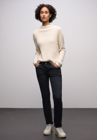 Kremowy sweter w dzianinę o luźnym kroju, zestawiony z ciemnymi dżinsami. Model nosi białe trampki z beżowymi podeszwami. Prosty, nowoczesny design.