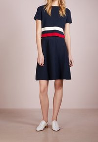 Marindress med en figursydd överdel och utställd kjol, med vita och röda randiga detaljer i midjan. Matcher med vita snörskor.