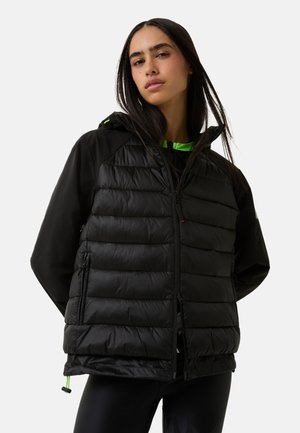 Frau in einer schwarzen, gepolsterten Jacke und schwarzen Leggings, stehend mit den Händen hinter dem Rücken vor einem weißen Hintergrund.