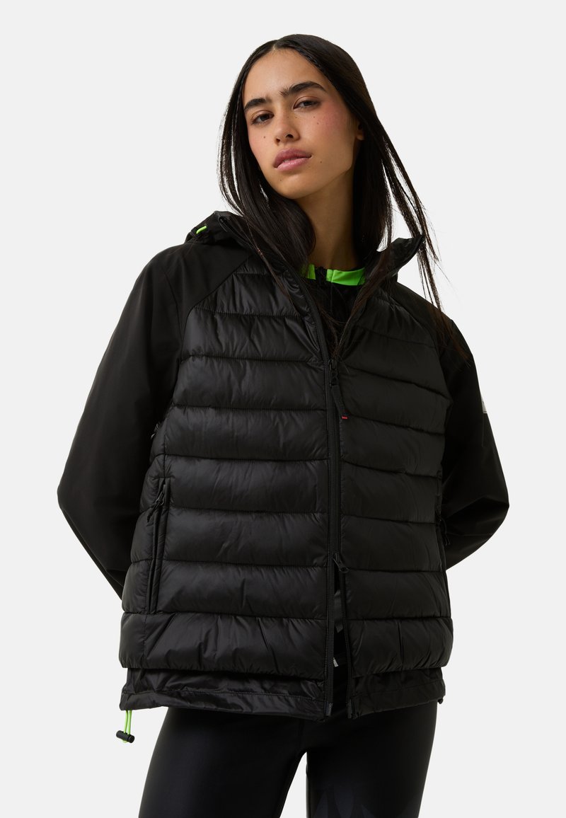 Kvinde iført en sort polstret jakke og sorte leggings, stående med hænderne bag ryggen mod en hvid baggrund.