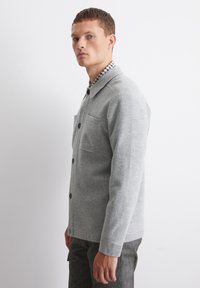 Grey button-up jacket in soft fabric with two chest pockets, featuring a plaid collar and black buttons. Tailored fit with long sleeves.