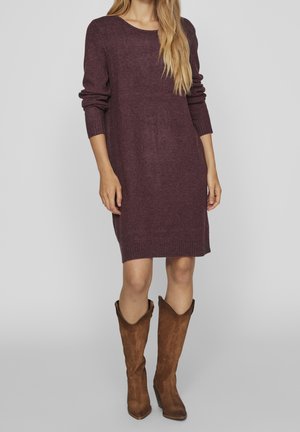 Vrouw die een paars, knielang trui-jurk met lange mouwen draagt, gecombineerd met middenkuithoog bruine suède laarzen, tegen een effen achtergrond.