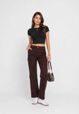 Donna che indossa una t-shirt nera corta, pantaloni cargo marrone scuro, sneakers bianche e tiene una borsa marrone scuro davanti a uno sfondo semplice.
