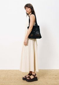 Schwarze gewebte Tasche mit strukturiertem Design und stabilen Trägern, kombiniert mit einem hellen gestreiften Kleid und dunklen Plateausandalen.