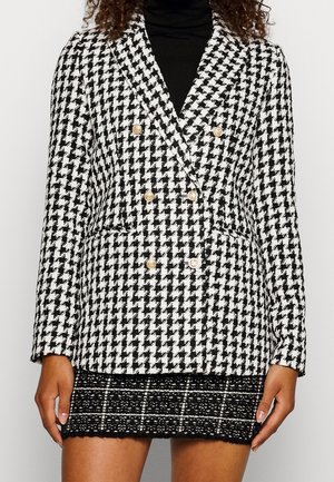 Frau trägt ein schwarz-weißes doppellagiges Hahnentrittmuster-Blazer mit goldenen Knöpfen über einem schwarzen Rollkragenpullover und einem schwarzen strukturierten Rock.