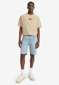 Beige cotton t-shirt with red logo patch, paired with light blue denim shorts. Worn with white socks and cream sneakers.