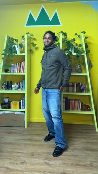 Sweat à capuche vert avec fermeture éclair et manches texturées, jean en denim et chaussures noires. En arrière-plan, des étagères remplies de livres et un mur jaune.