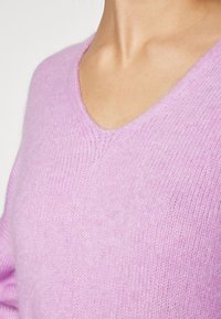 Femme portant un pull violet doux à col en V montrant la texture et le détail du décolleté sur un fond uni.