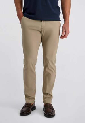 Mann trägt beige Slim-Fit-Hosen, dunkelbraune Loafers und ein marineblaues Hemd, steht mit entspannten Händen an den Seiten.