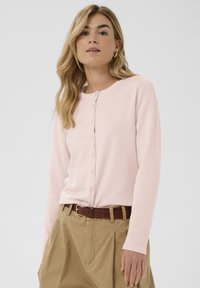 Svetlo roza jopica z dolgimi rokavi in okroglo izrezom, s sprednjim zapenjanjem na gumbe in mehko teksturo, kombinirana z bež hlačami in rjavim pasom.