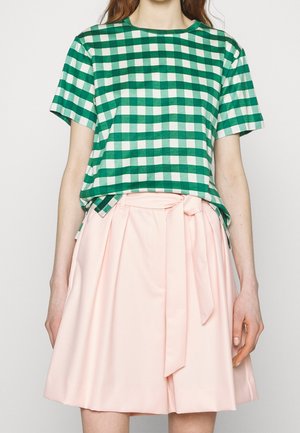 Mujer con camisa de manga corta de cuadros verdes y blancos metida en una falda de talle alto de color rosa pálido con cinturón anudado.