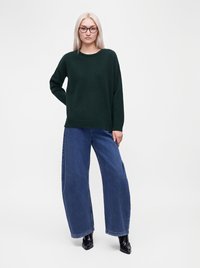 Femme aux longs cheveux blonds et lunettes, portant un pull vert foncé, un jean bleu à jambes larges et des bottines noires, debout sur fond blanc.