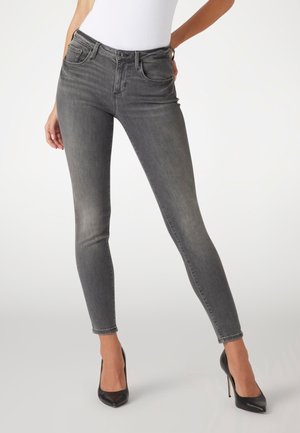 Femme portant un jean skinny gris moulant et des chaussures à talons noirs, debout devant un fond blanc uni.