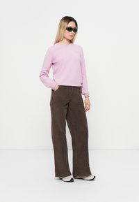 Lys rosa strikket genser, brune kordfløyelsbukser med vid ben, og svarte solbriller. Føttene i hvite sneakers med svarte detaljer.