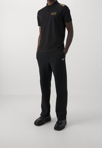 Man in een zwart EA7 poloshirt met gouden logo en strepen, zwarte broek met wit logo, en zwarte instappers, staand met de handen in zijn zakken.