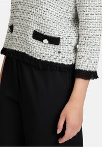 Weiß-schwarze Tweedjacke mit strukturiertem Finish, ausgestattet mit zwei schwarzen Taschen, silbernen Knöpfen und Fransen am Saum.