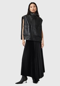 Gilet in pelle nera senza maniche, con tasche frontali e colletto alto; abbinato a una gonna nera plissettata e stivali neri a punta.
