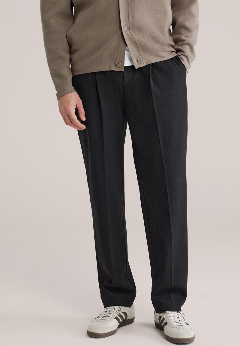 Pantalon gris foncé sur mesure avec un devant plissé, associé à un cardigan marron clair boutonné et des baskets blanches avec des accents verts.