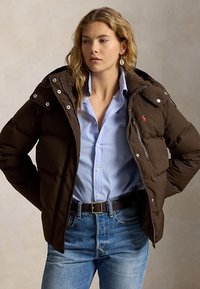 Femme portant une doudoune marron sur une chemise bleu clair boutonnée et un jean bleu, debout les mains sur les hanches devant un fond uni.