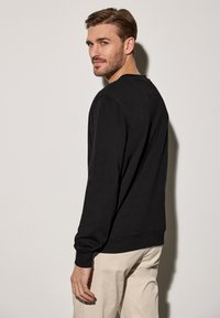 Svart bomullströja med långa ärmar, rund halsringning och ribbade ärmslut. Modellen står sidledes mot en neutral bakgrund.