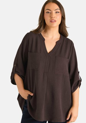 Vrouw die een loszittende donkerbruine blouse met V-hals, opgerolde mouwen en opgestikte zakken draagt, met één hand in haar zak, staand tegen een effen achtergrond.