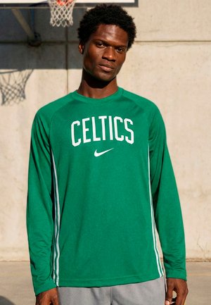 Muž oblečený v zelenom dlhoročnom tričku Celtics stojí vonku blízko basketbalového koša a betónovej steny.