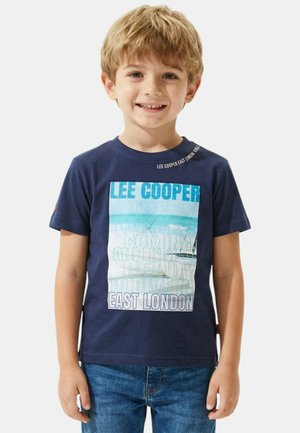 Jeune garçon souriant aux cheveux blonds portant un t-shirt Lee Cooper bleu marine et un jean bleu, debout devant un fond blanc.