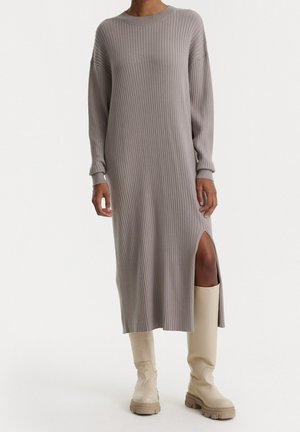 Geripptes, langärmliges Kleid in Hellgrau mit einem seitlichen Schlitz und lockerer Passform. Kombiniert mit beigen, knielangen Stiefeln.