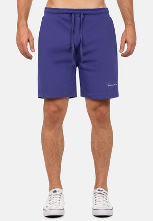 Hombre con pantalones cortos morados con cordón y zapatillas blancas, de pie frente a un fondo claro y sencillo.
