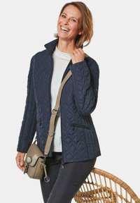 Navy gesteppte Jacke mit Stehkragen, mit Reißverschlusstaschen, strukturiertem Stoff, über einem weißen Oberteil getragen, kombiniert mit einer kleinen Umhängetasche.