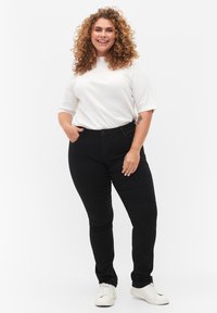 Black fitted jeans with a smooth texture, paired with a white ribbed top and white sneakers. The model has curly hair and hands in pockets.
