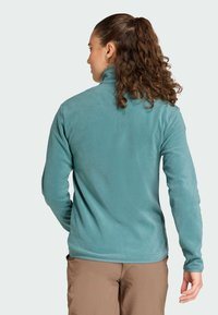 Giacca in fleece turchese con colletto alto e maniche lunghe, texture liscia, design aderente, abbinata a pantaloni marrone chiaro.