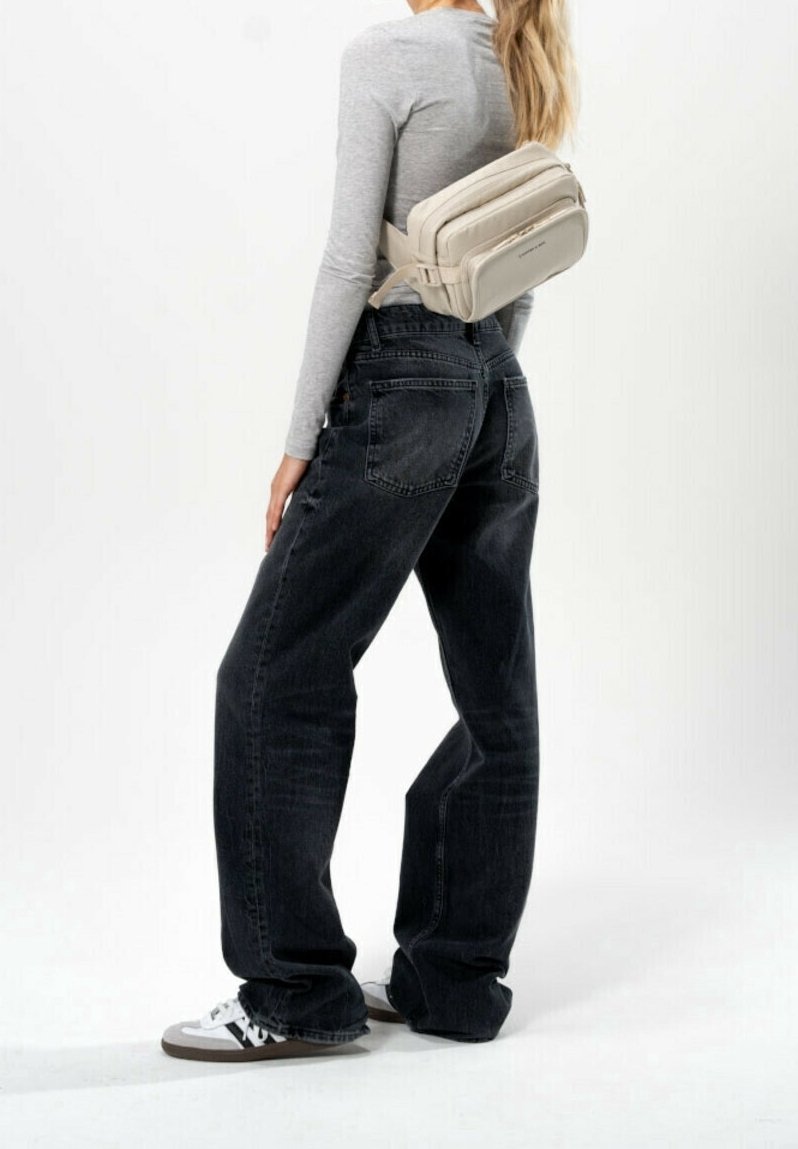 Personne portant un jean noir à jambes larges, un haut gris à manches longues, des baskets blanches et un sac bandoulière beige sur un fond blanc uni.
