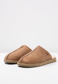 Brown suede slip-on slippers with a soft fur interior, rounded toe design, and a textured sole for comfort. Small branding label present.
