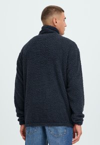 Pullover in pile navy con colletto alto, tessuto strutturato, vestibilità ampia e polsini a coste, indossato con jeans azzurri chiari.