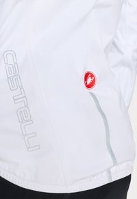 Weißer Fahrradjacke aus leichtem Material. Besitzt ein graues Logo, ein rotes Logo-Abzeichen und reflektierende Akzente an der Seite.