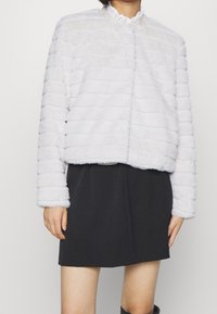 Bílá umělá kožešinová bunda s texturovaným pruhovaným vzorem a vysokým límcem, v kombinaci s černou sukní s uvolněným, zkráceným střihem.