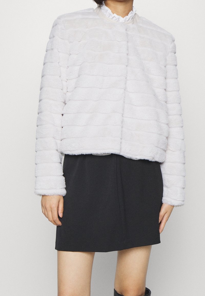 Bílá umělá kožešinová bunda s texturovaným pruhovaným vzorem a vysokým límcem, v kombinaci s černou sukní s uvolněným, zkráceným střihem.