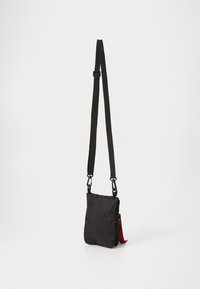 Borsa a tracolla nera realizzata in tessuto texturizzato, con una tracolla regolabile e un'etichetta rossa in contrasto sul lato. Forma compatta e rettangolare.