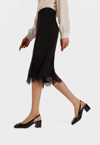 Falda de satén negra con dobladillo de encaje y aberturas, combinada con zapatos de charol negros tipo slingback con tacón ancho y hebilla decorativa.